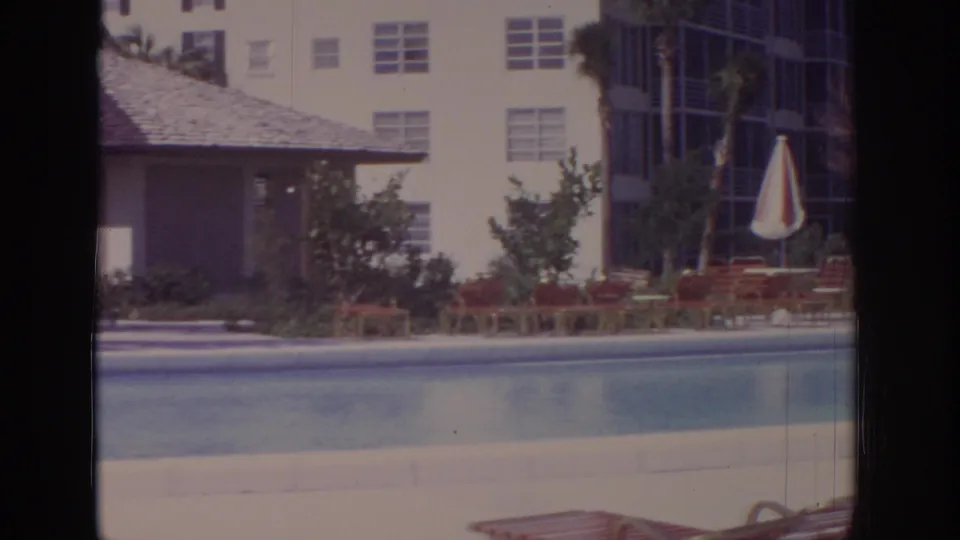 1973 Naples, Florida pool area with calm water, an umbrella, lounge chairs, a screened structure, landscaping, and apartment windows.