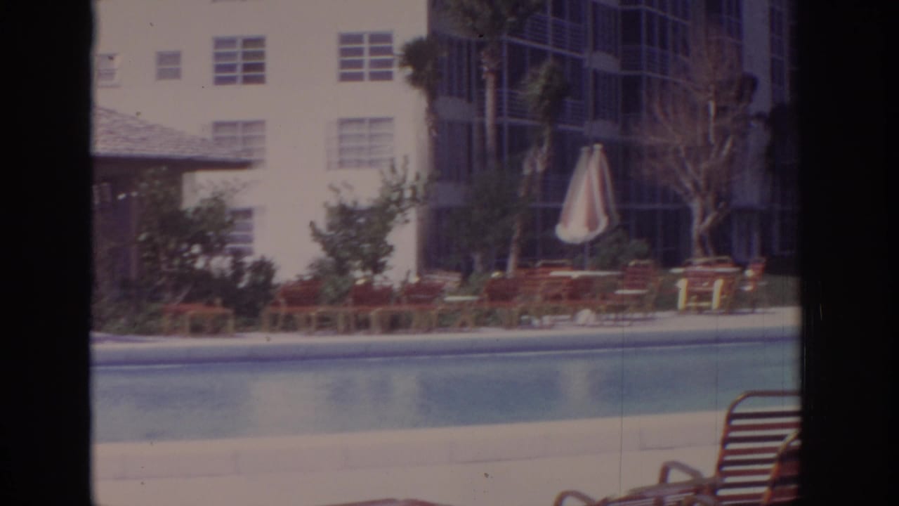 Hotel swimming pool with lounge chairs, striped umbrella, and multi-story buildings behind it.
