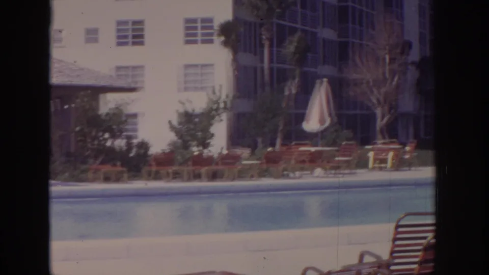 1973 Naples, Florida hotel pool area with red lounge chairs, a closed striped umbrella, palms, and modern resort buildings.
