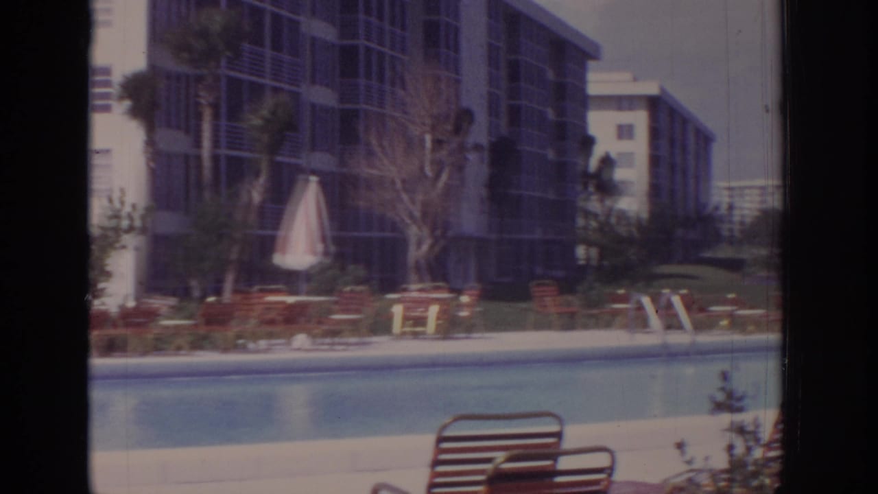 Pool deck with lounge chairs, striped umbrella, trees, and dark-screened hotel balconies.