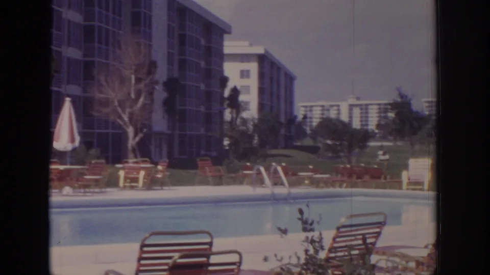 1973 Naples hotel pool view showing a pool ladder, rows of red lounge chairs, tropical shrubs, and resort buildings beyond.