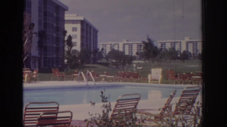 Wide 1973 Naples, Florida poolside frame with lounge chairs in the foreground, hotel grounds, trees, and distant apartment blocks.