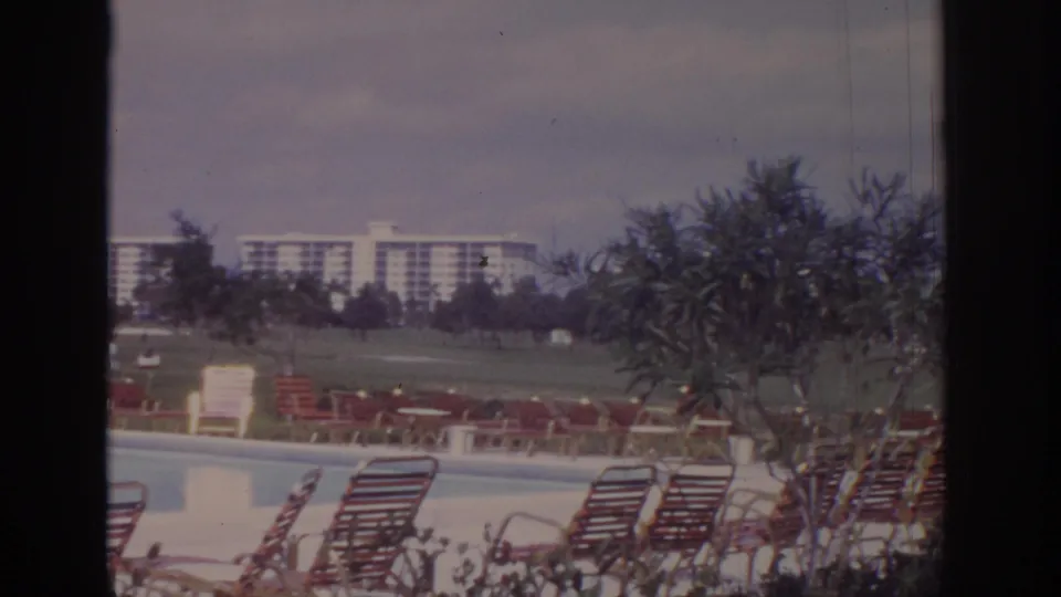 1973 resort pool area in Naples, Florida, showing lounge chairs, trees, lawn, and distant modernist buildings on 8mm film.
