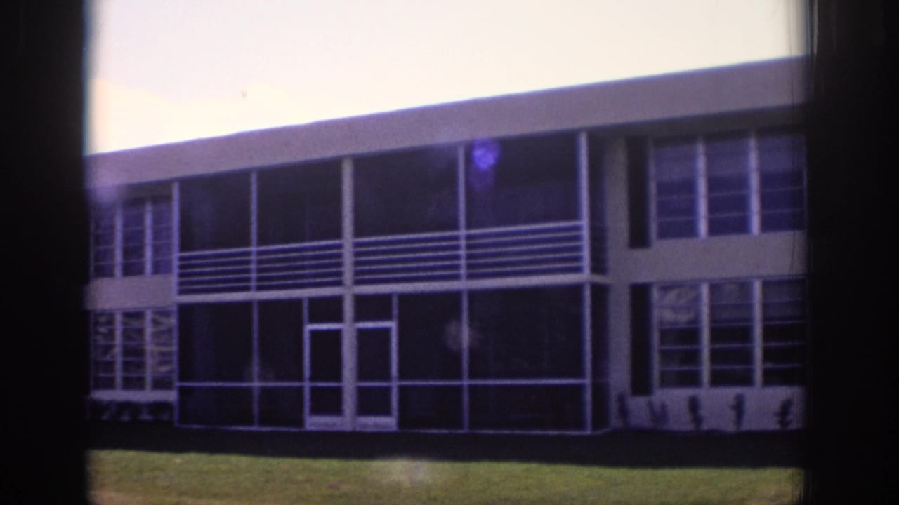 Two-story modernist building with glass panels, white railings, lawn, and bright sky.