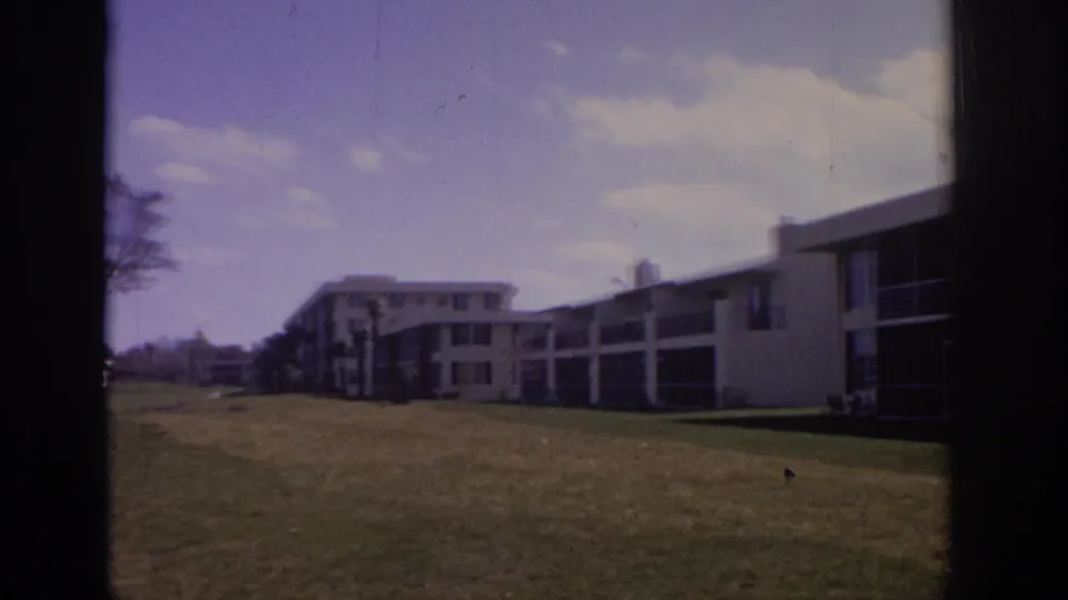 Soft 8mm frame from Naples in 1973 showing a lawn, white apartment blocks, screened porches, and a tree near the edge.