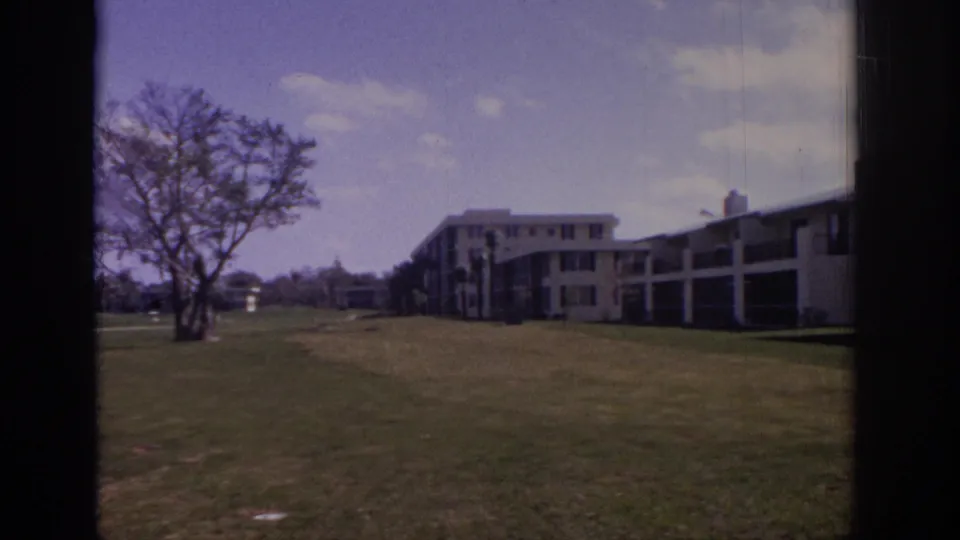 1973 Naples, Florida home-movie frame with a solitary tree, wide lawn, and modern white apartment buildings in the background.