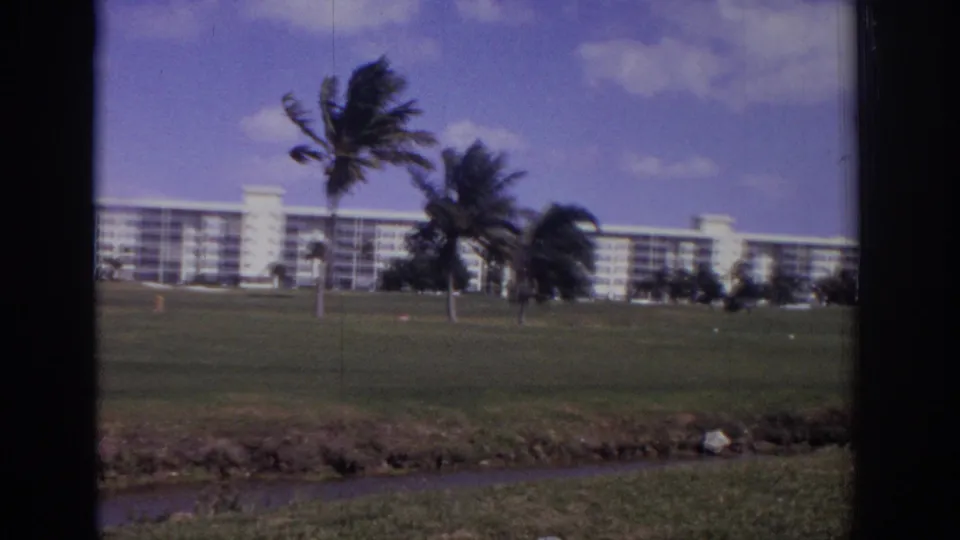 1973 Naples, Florida home-movie frame with palms, white multi-story buildings, lawn, and a narrow foreground water channel.
