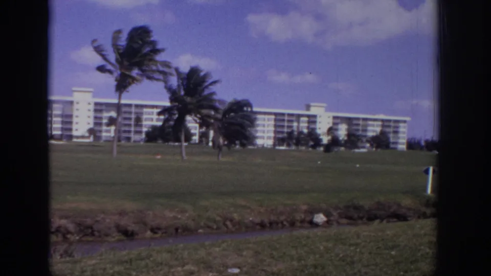 1973 Naples, Florida resort grounds with a white hotel, wind-bent palms, grass, and dark window edges in 8mm footage.
