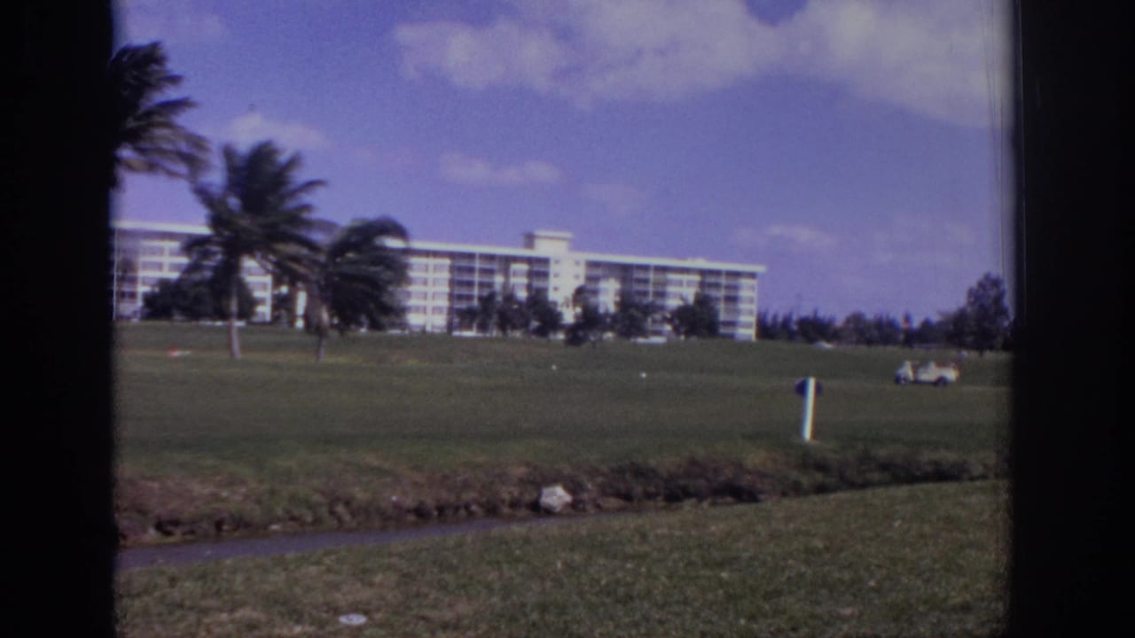 Resort building, palms, golf-course grass, and a small distant cart under a blue sky.
