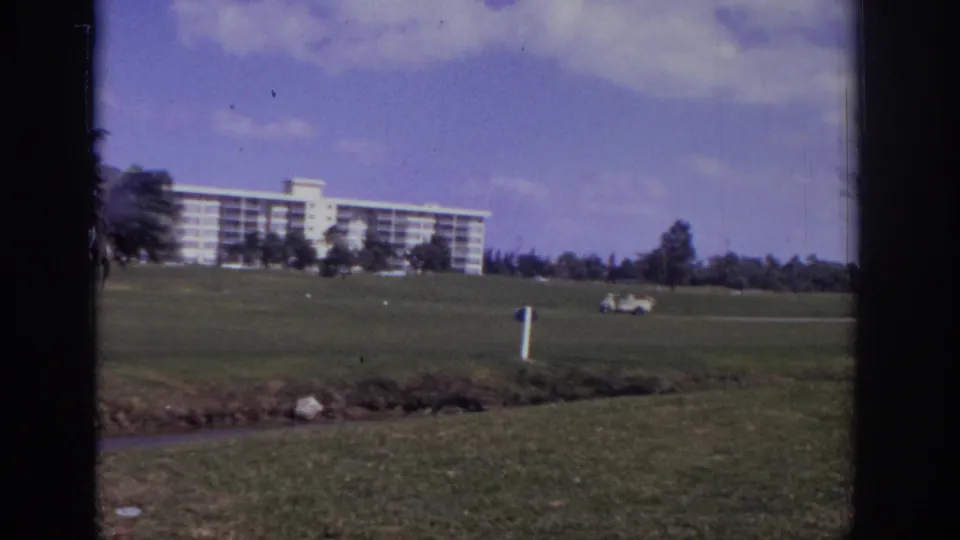 1973 Naples, Florida home-movie frame showing a white resort across green grounds, with a distant golf cart and soft 8mm color.