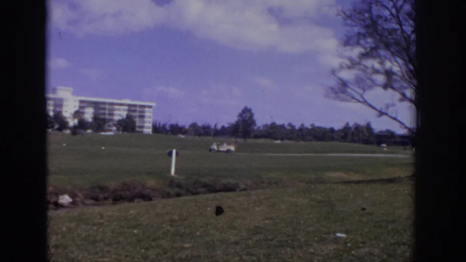 1973 Naples, Florida resort exterior with green course, distant cart, white hotel, clouds, and tree branches along the frame edge.
