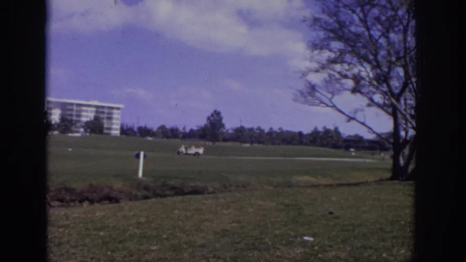 1973 Naples, Florida golf-course view with a distant resort-style building, golf cart, open lawn, and dark 8mm frame edges.