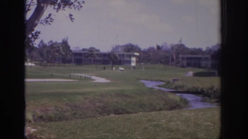 Wide 1973 Naples golf course shot showing a water channel, fairway, distant buildings, trees, and small golfers across the lawn.