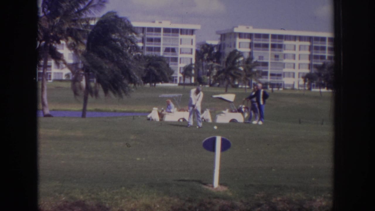 Men on a golf course near a white cart, with palms and white apartment buildings behind them.