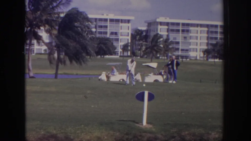 1973 Naples, Florida home-movie frame showing men golfing on a green course with a white cart, palms, and condominium-style buildings in the background.