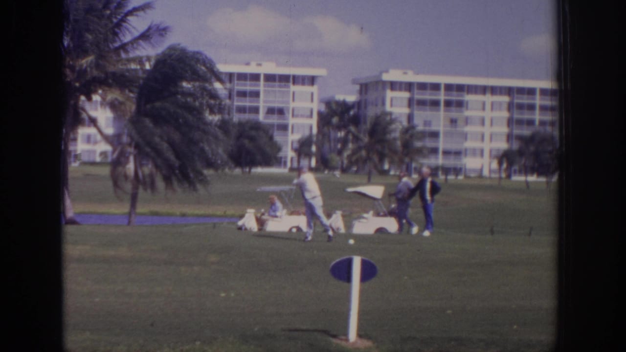 A golfer stands near a white cart while two other men wait on the course.