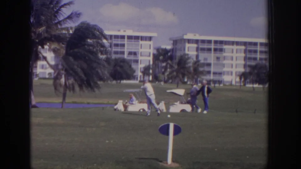 1973 Naples, Florida frame with a golfer near a white cart, other men nearby, palm trees, and white residential-style buildings beyond the green.