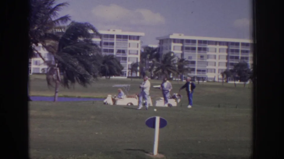 1973 Naples, Florida golf scene showing several men beside a white cart on a green course, with palms and multi-story buildings in the distance.