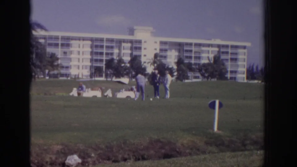 1973 Naples, Florida frame showing distant golfers, a white cart, palms, and a large white condominium-style building across the course.