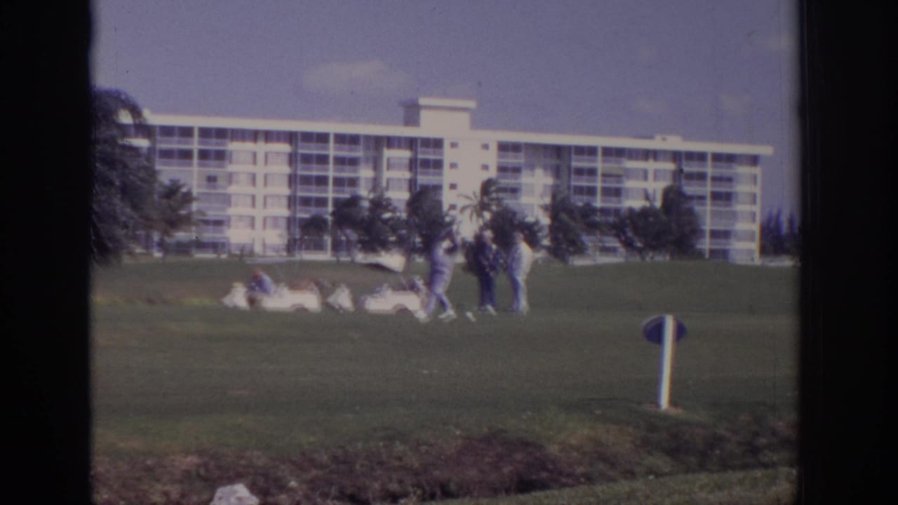 Golfers on a green seen through a dark window frame with a white resort building behind them.