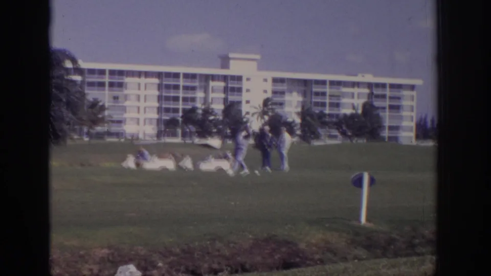 1973 Naples, Florida resort golf scene viewed through a window, with small figures on the green and a white hotel building in the background.
