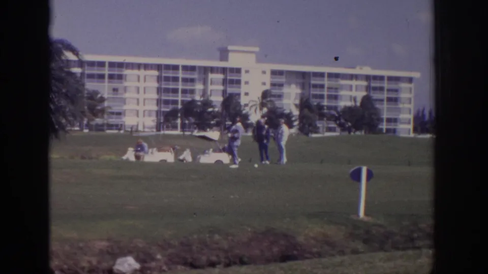 Small figures gather on a Naples resort golf green in 1973, filmed through a window with palms and a white hotel behind them.