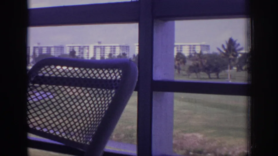 The foreground mesh chair fills much of the frame while a Naples balcony view shows palms, grass, and distant buildings outside.