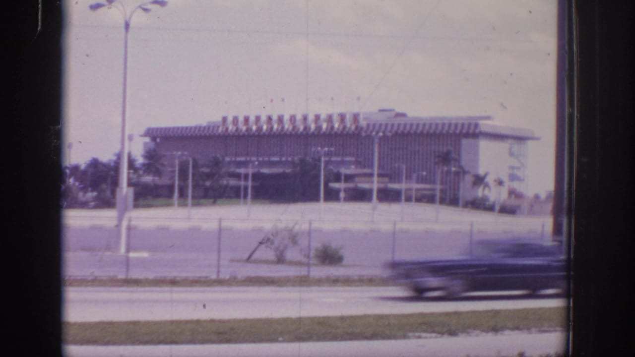1973 Naples, Florida 8mm frame showing a large striped-roof building across a road, with chain-link fence, poles, and a passing car blur.