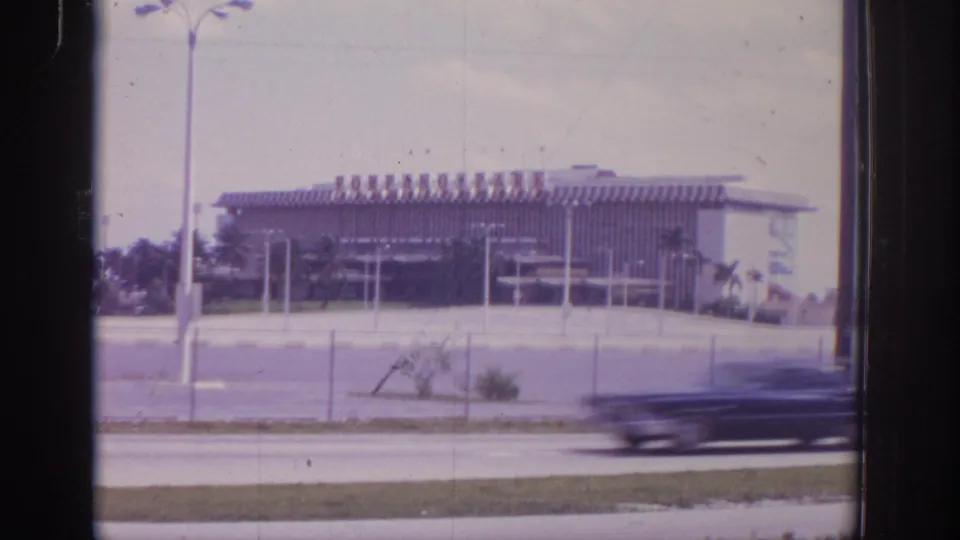 1973 Naples, Florida 8mm frame showing a large striped-roof building across a road, with chain-link fence, poles, and a passing car blur.