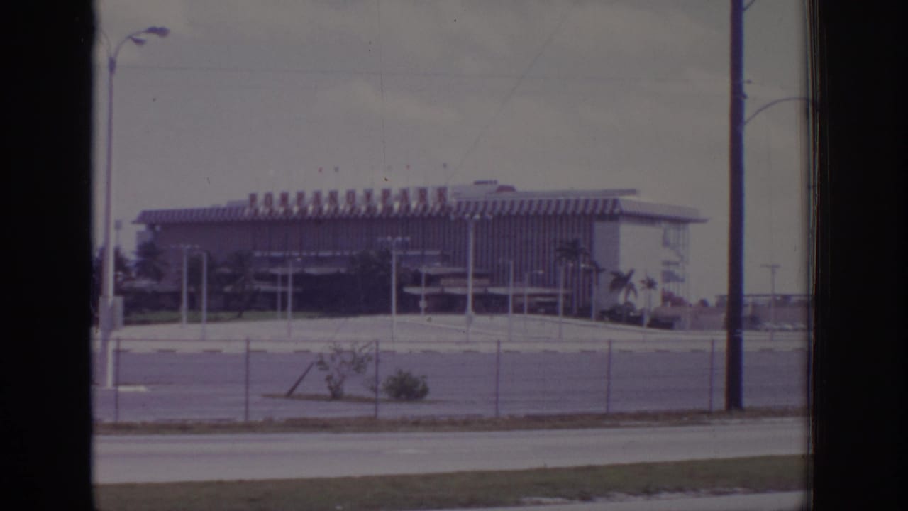 1973 Naples, Florida home-movie frame of a large roadside building viewed through film gate edges, with fence, poles, and palm landscaping.