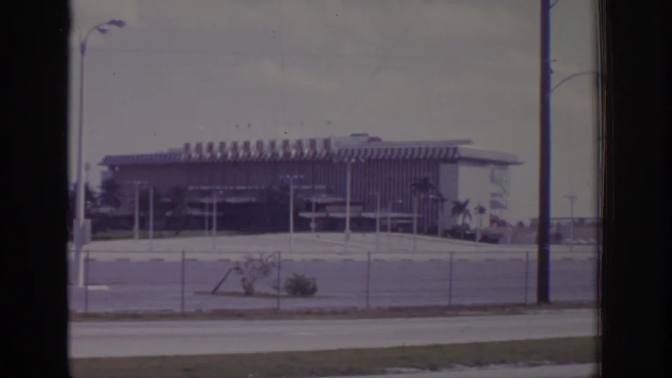 1973 Naples, Florida home-movie frame of a large roadside building viewed through film gate edges, with fence, poles, and palm landscaping.