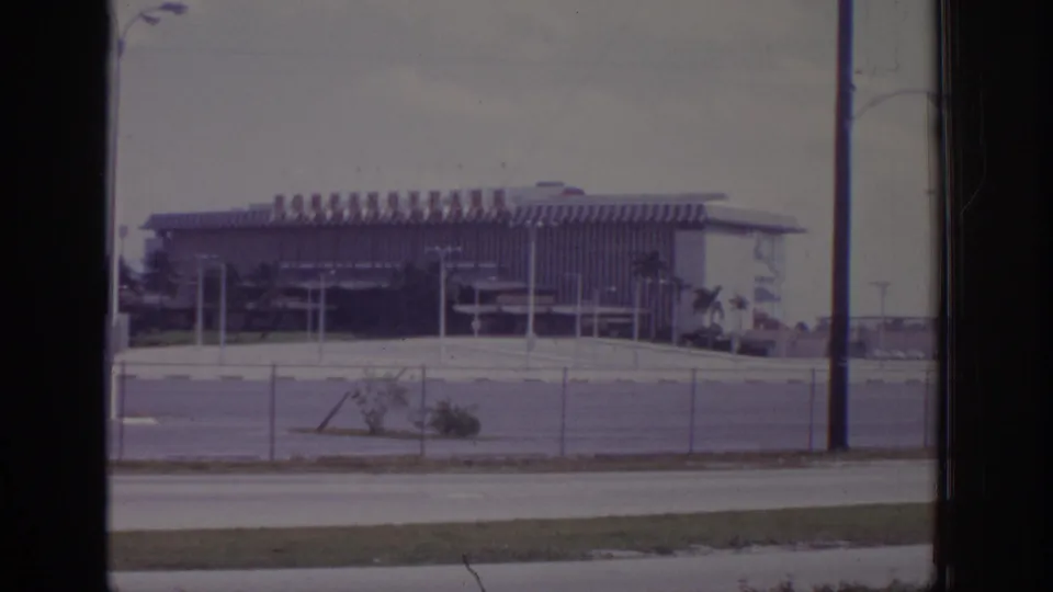 1973 Naples, Florida frame showing the same fenced roadside building, with a metal mesh chair partly visible from the filming position.