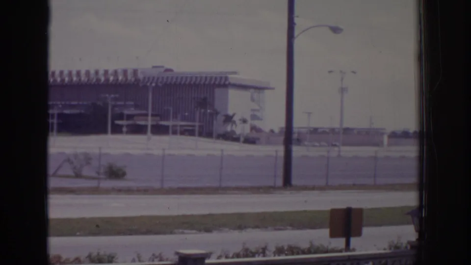 1973 Naples, Florida 8mm frame with a striped-roof roadside building beyond a chain-link fence, roadside poles, grass, and a small sign.