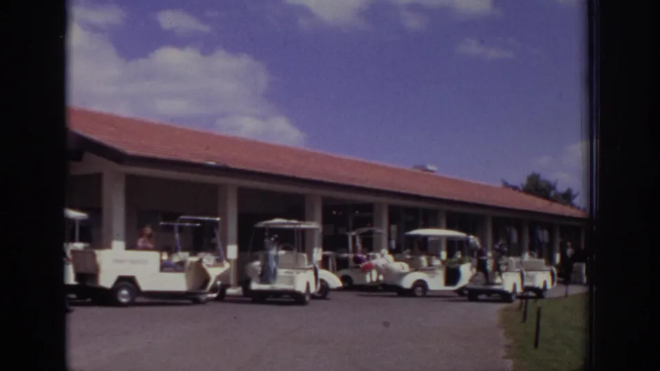1973 Naples, Florida frame showing white golf carts, people, a red-roofed course building, and a strip of grass beside the paved area.