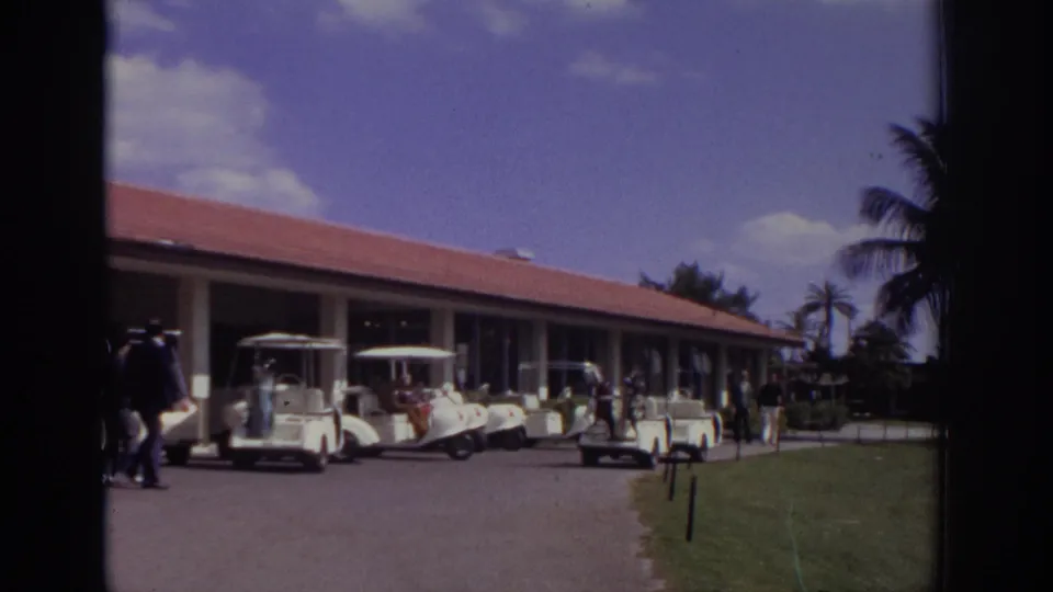 1973 Naples, Florida home-movie frame with golf carts near a red-roofed building, people on the drive, lawn, and palm trees in the background.