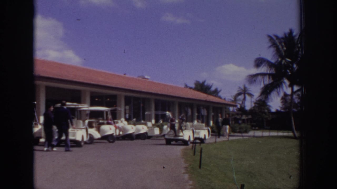Golf carts parked beside a low clubhouse while adults stand nearby on a sunny resort course.