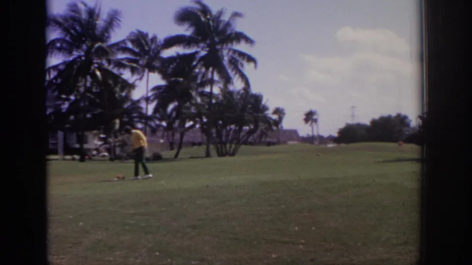 1973 Naples, Florida resort golf course with a golfer in yellow, nearby cart, wind-blown palms, fairway, and open sky.