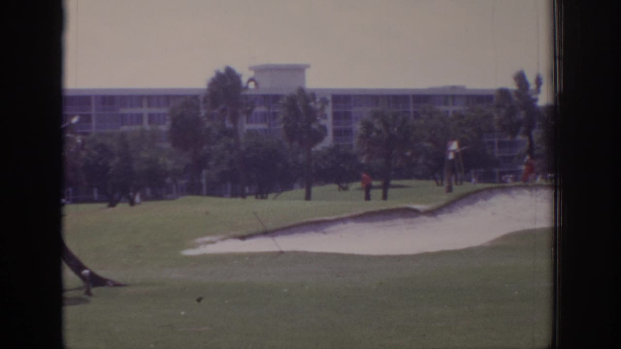 Golf course bunker and green with palms, distant people, and a large building beyond the fairway.