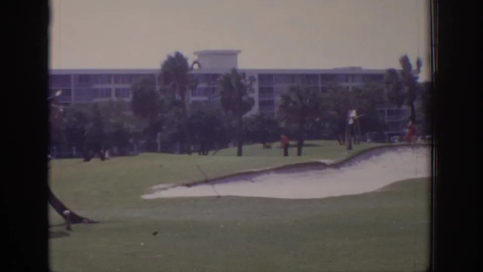 1973 Naples, Florida home-movie frame of a golf course sand trap and green with palms moving in wind, distant people on the course, and a resort-style building in the background.