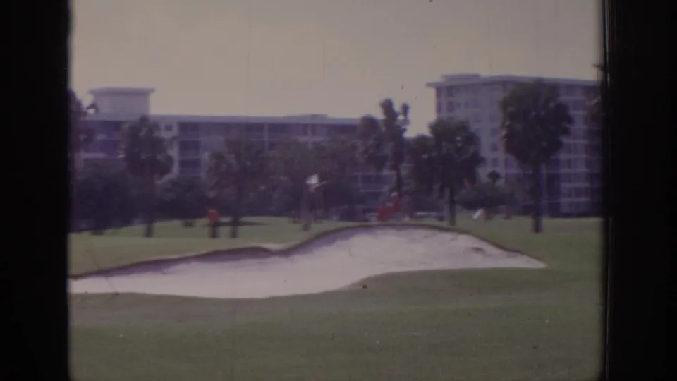 1973 Naples, Florida frame showing a wide view across a golf course bunker toward palms, distant figures, and multi-story resort-style buildings under windy conditions.