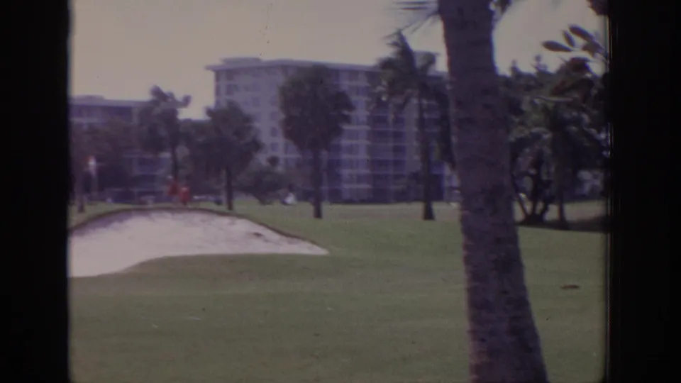 1973 Naples, Florida 8mm frame with a palm trunk close to the camera, a sand bunker and green beyond it, and resort-style buildings partially obscured by trees.