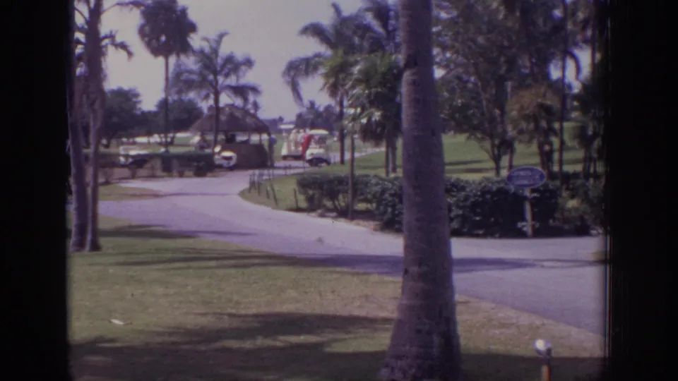 1973 Naples, Florida home-movie frame of a palm-lined resort golf course path, distant golfers, shrubs, and a small sign in the foreground.