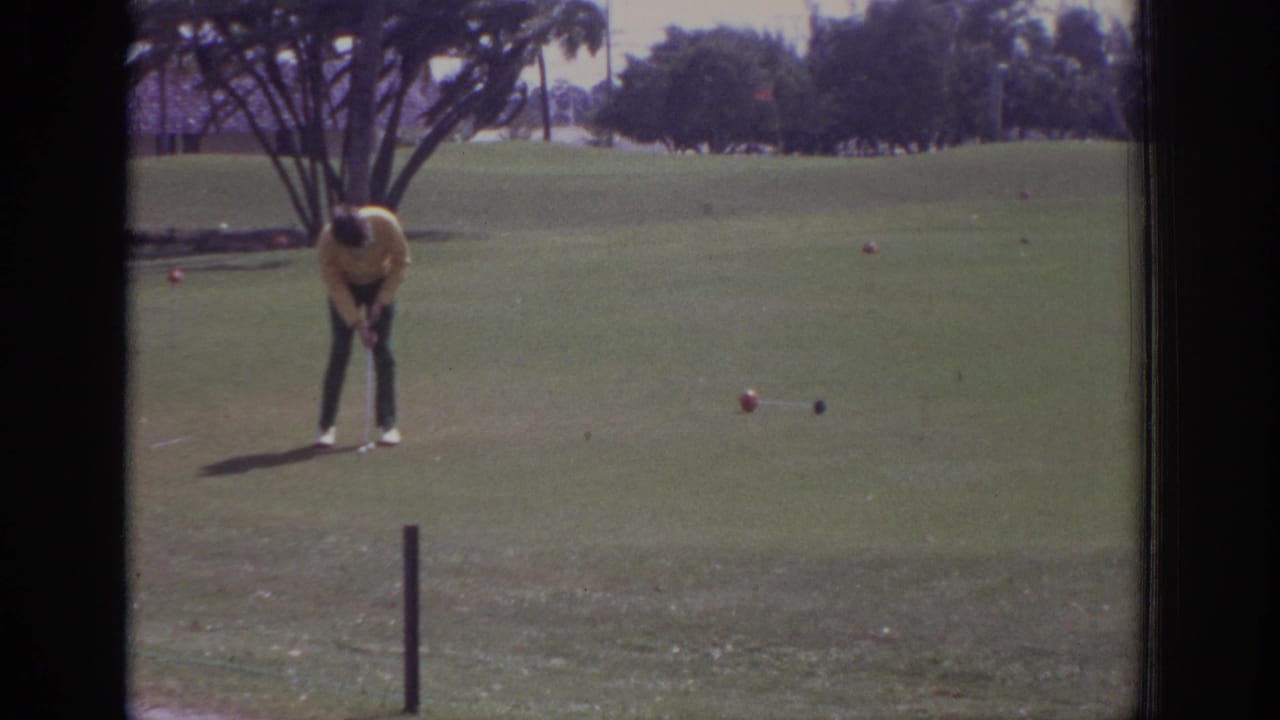 Golfer in a yellow shirt bends over near a ball on a green golf course.
