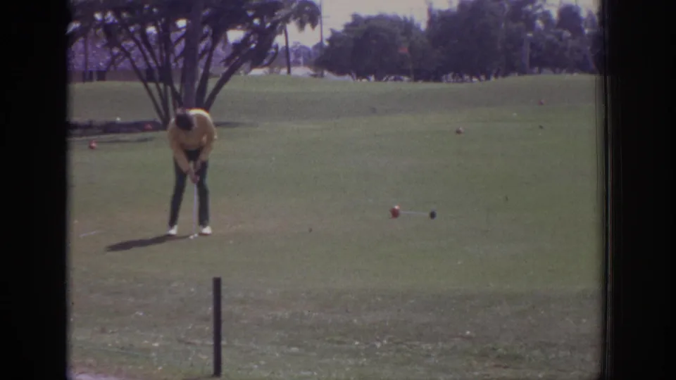 In supplied 1973 Naples, Florida footage, a golfer in a yellow shirt prepares near a ball on a sunny course with trees behind him.