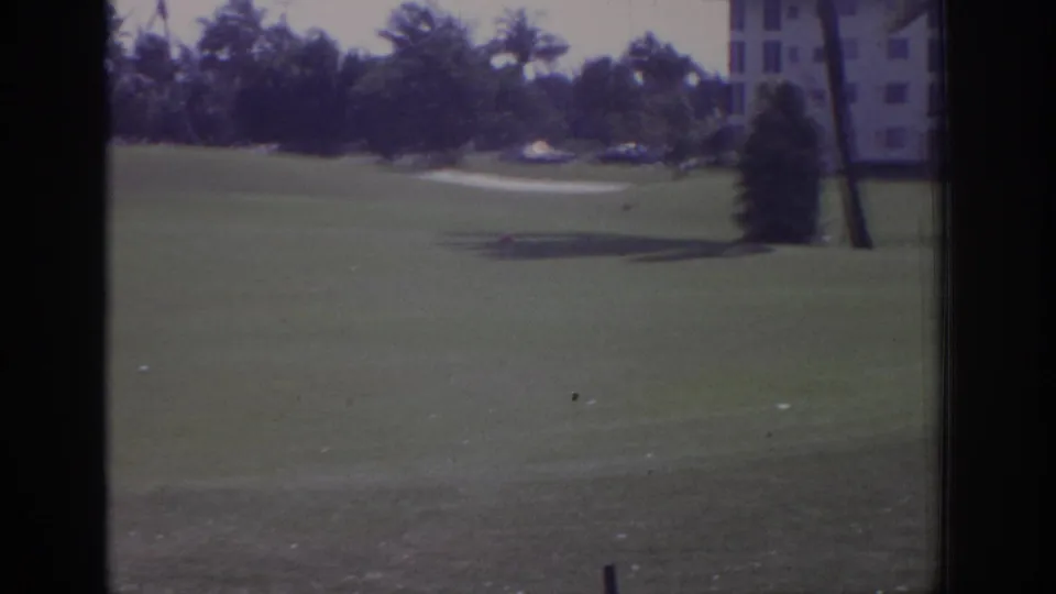 The final frame looks across the Naples course toward a pale path, palms, a small shrub, and a distant building at the right edge.