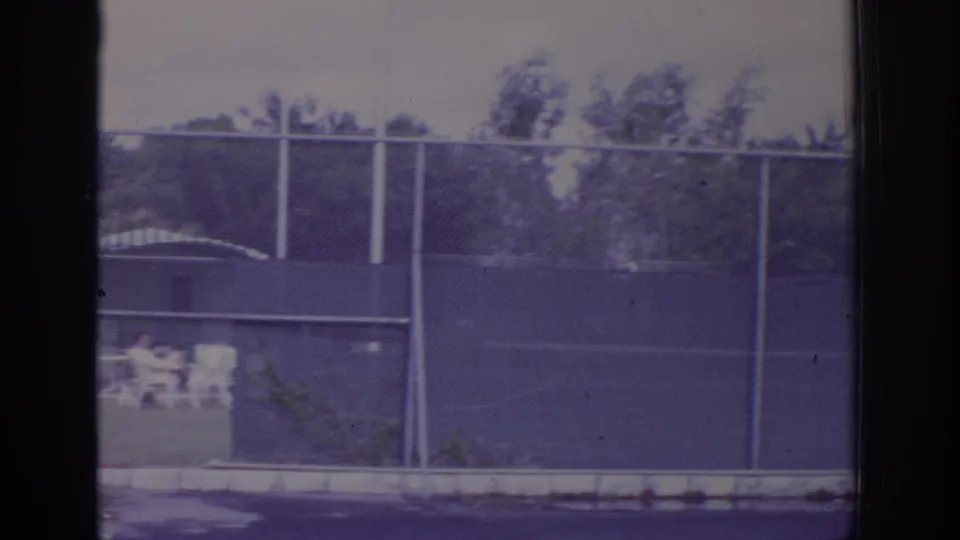 1973 Naples, Florida 8mm frame of a fenced racquet club area with chairs, trees, and indistinct people in the background.