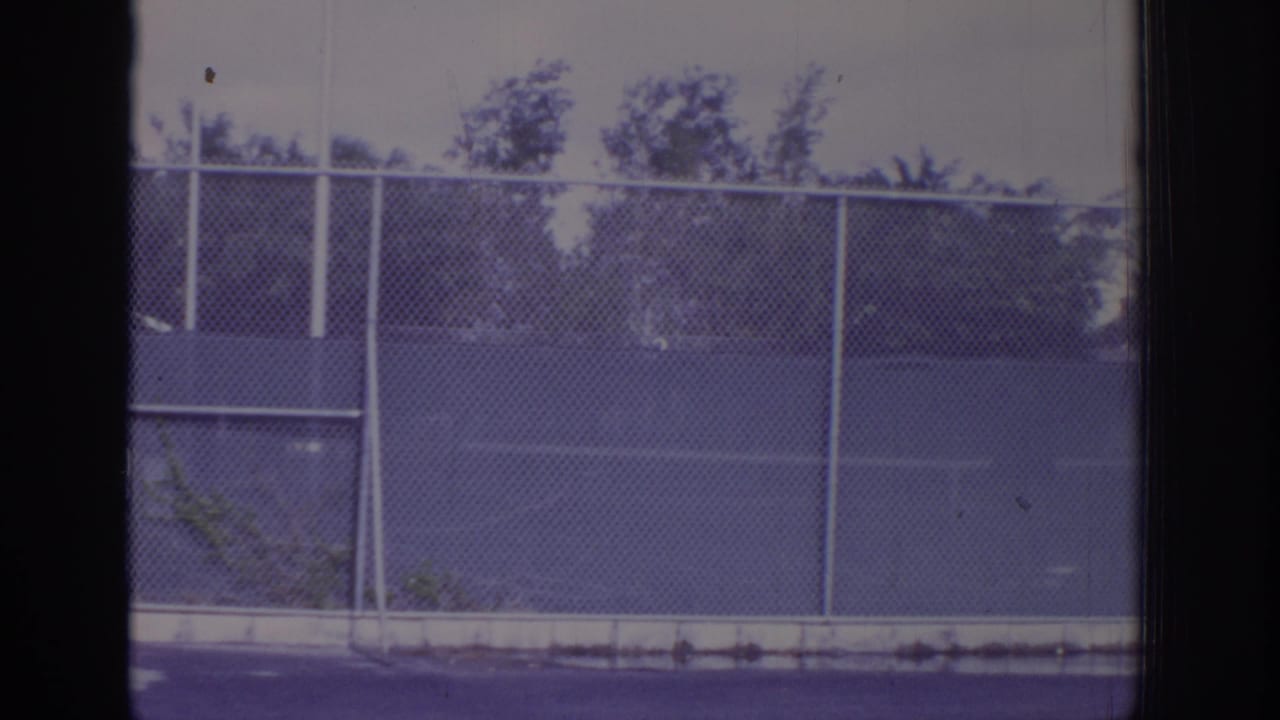 Chain-link fence beside outdoor tennis courts seen through dark film-frame edges.