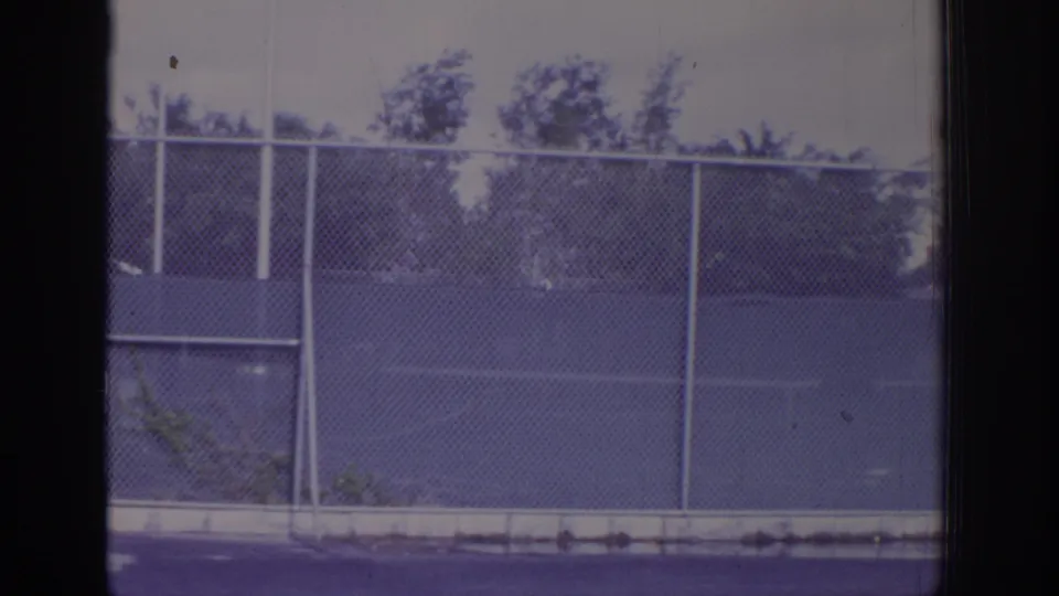 1973 Naples, Florida home-movie frame showing chain-link fencing beside outdoor tennis courts, with trees beyond and dark frame edges.