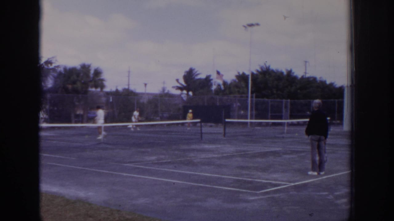 Outdoor tennis court with several adult players, net, fencing, trees, and flag in the background.