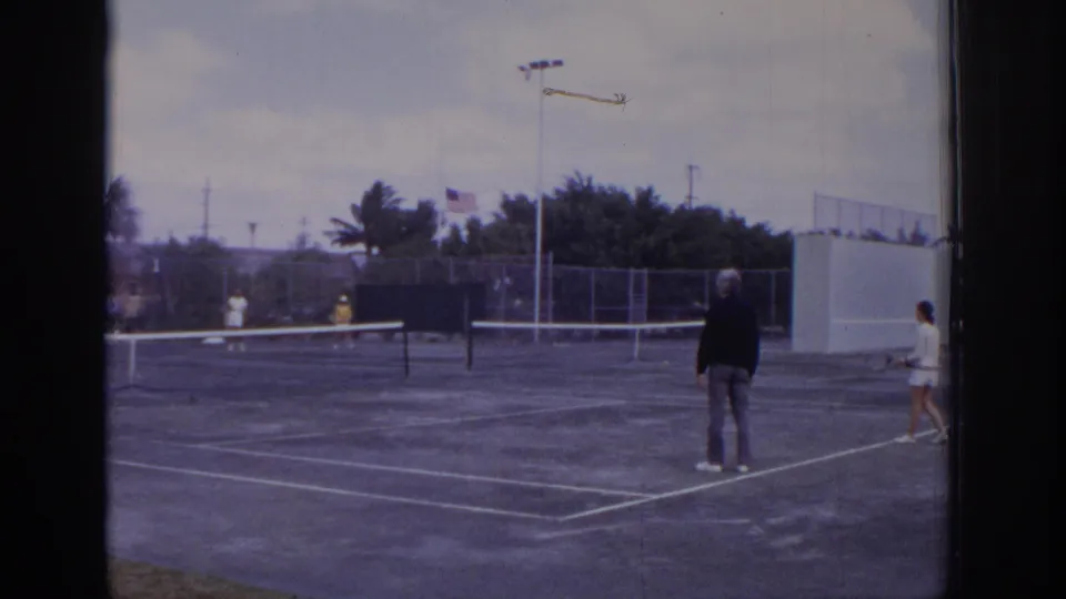 1973 Naples, Florida frame showing mixed doubles tennis on an outdoor court, with a foreground male player and other players across the net.